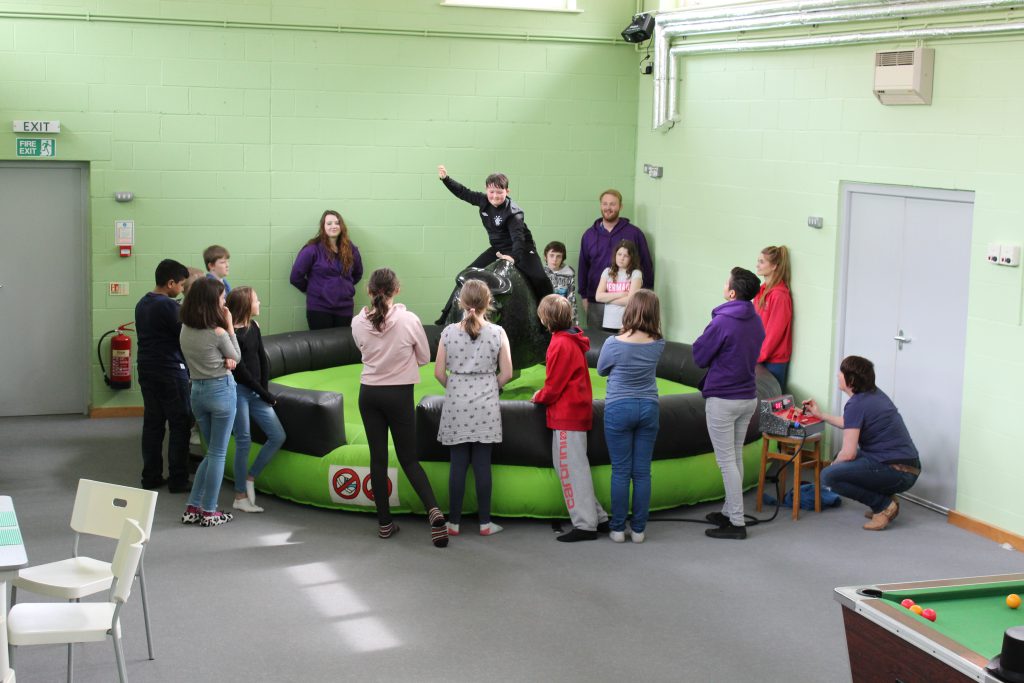 A group of children are watching a boy on a mechanical bucking bronco in a large games room.