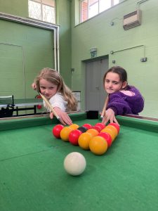 Two children are poised to play pool on a green pool table