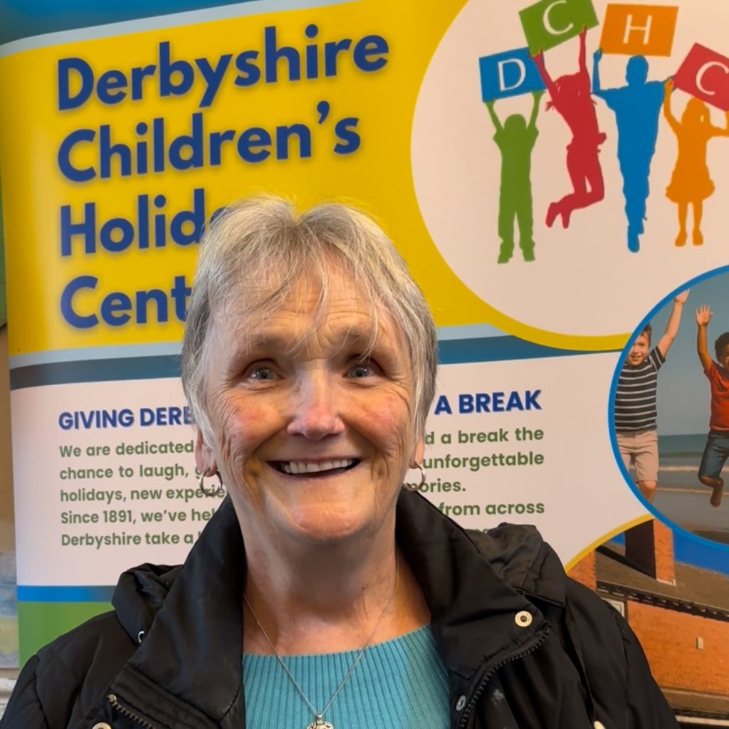 A lady with short grey hear, a blue jumper and a smiley face is standing in front of a colourful DCHC banner speaking to the camera.